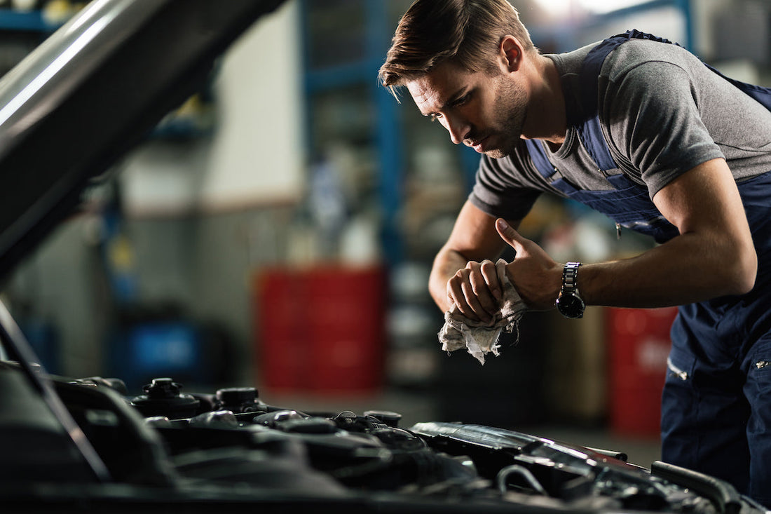 A mechanic in blue overalls wipes a car engine component with a white cloth in comprehensive battery care kits.