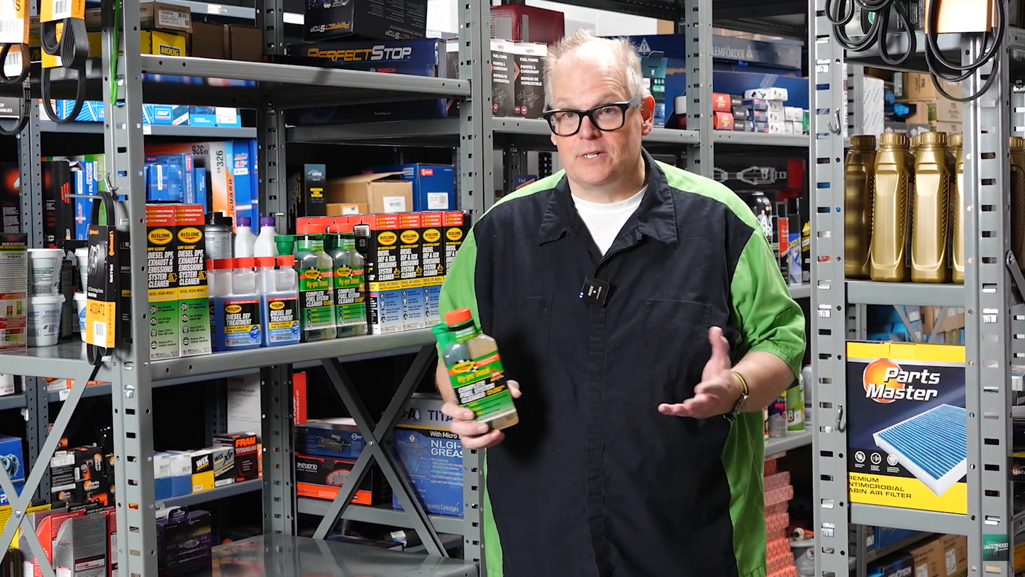 Man holding a bottle of Rislone Hy Per Complete Fuel System Treatemnt for Diesel, in a warehouse setting with shelves stocked with products.